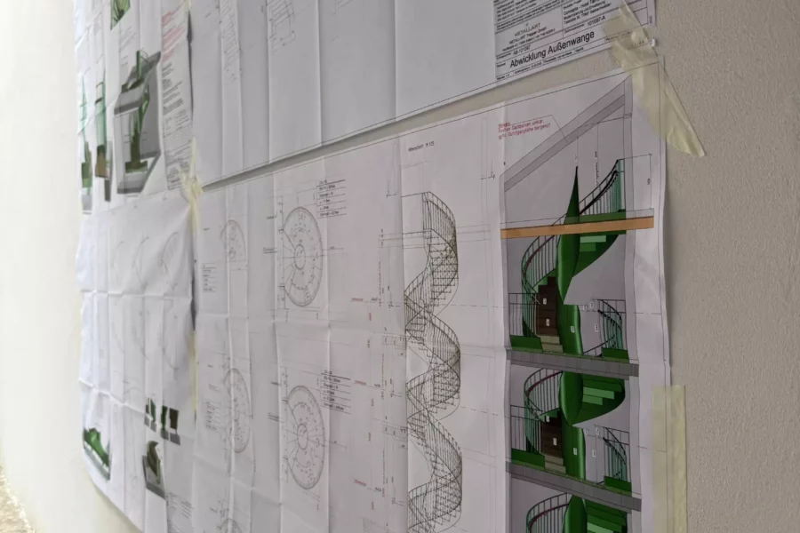 Architekturpläne einer grünen Wendeltreppe, an eine weiße Wand im Rohbau geklebt, Detailzeichnungen und 3D-Ansichten sichtbar