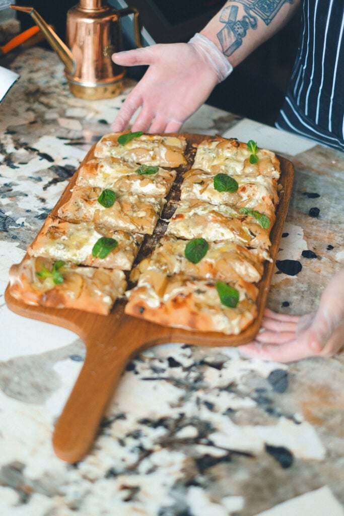 Flammkuchen mit Käse und Kräutern auf Holzbrett präsentiert von Person mit Handschuhen in Boutique Resort Küche Schwarzwald