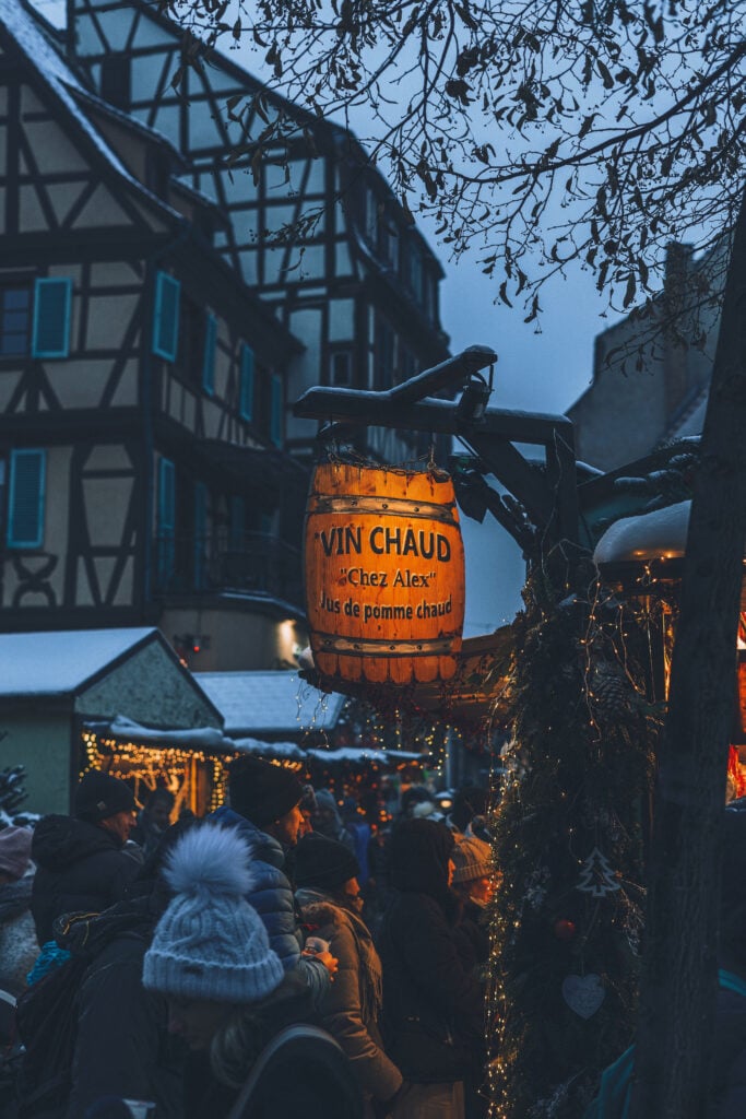 Weihnachtsmarkt mit beleuchtetem Schild Vin Chaud und Menschen vor Fachwerkhäusern im Schwarzwald bei Dämmerung