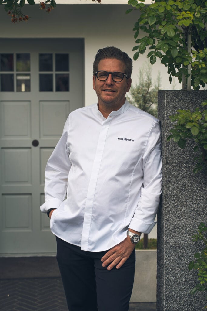 Mann mit Brille und weißer Kochjacke lehnt an Steinmauer vor Tür, Außenbereich mit Pflanzen, Boutique Resort Schwarzwald