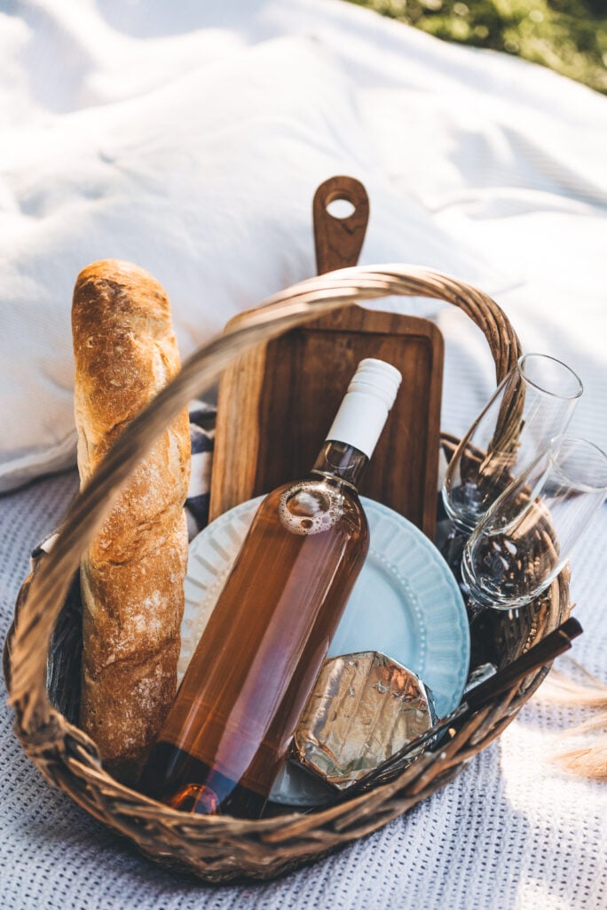 Picknickkorb mit Baguette, Flasche Roséwein, zwei Gläsern, Käse und Holzbrett auf weißer Decke im Freien im Schwarzwald